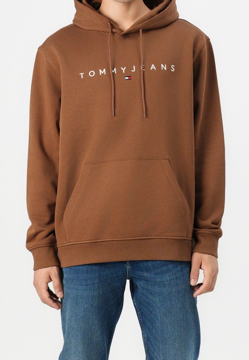 Homme portant un sweat à capuche marron avec une poche avant et un logo "Tommy Jeans" au-dessus d'un petit drapeau, associé à un jean bleu.