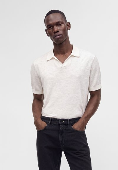 Homme portant une chemise blanche à manches courtes avec col et un pantalon noir, debout les mains dans les poches devant un fond uni.