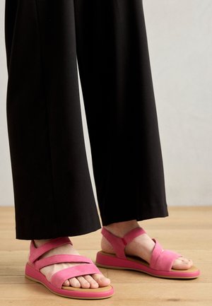 Lyserøde ruskinds sandaler med krydsede stropper og en tykk, polstret sål. Båret med sorte brede bukser, som afslører tæer og svangen.