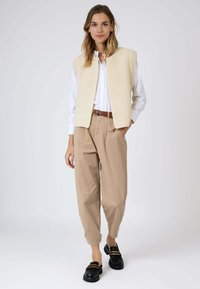 Gilet en tricot crème porté sur une chemise blanche à boutons, associé à un pantalon beige et des mocassins noirs, avec un motif texturé et une coupe décontractée.