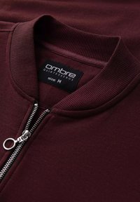 Bourgogne zip-sweater med ribbet krave, sølv lynlås og mærkeetiket "ombre QUINTESSENCE" der angiver størrelse M. Blød tekstur.