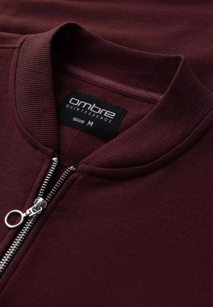 Bordeaux zip-up sweater met geribbelde kraag, zilveren ritsbeslag en merklabel "ombre QUINTESSENCE" dat maat M aangeeft. Zachte textuur.