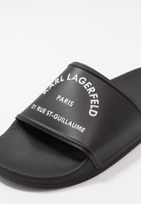 Juodos slystančios sandalai su tekstūruotu matinio paviršiumi. Yra ryškus baltas ženklas: "KARL LAGERFELD PARIS 21 RUE ST-GUILLAUME."