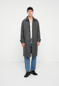 Grijze wollen overjas met een kraag, lange mouwen en knoopsluiting, gedragen over een witte tanktop en blauwe jeans. Zwarte schoenen maken de look compleet.