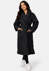 Zwarte lange gewatteerde robe met een brede kraag, een ceintuur in de taille en twee voorzakken, gemaakt van zachte, gladde stof; gecombineerd met witte sneakers.