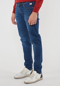 Uomo che indossa jeans blu e sneakers bianche, in piedi con le mani rilassate ai lati su uno sfondo bianco semplice.