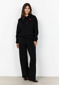 Felpa nera con un piccolo cuore rosso, abbinata a pantaloni neri a gamba larga. Entrambi i capi presentano una texture morbida e una vestibilità rilassata.