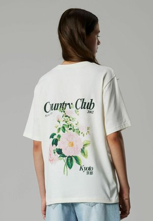 Donna con lunghi capelli castani che indossa una T-shirt bianca oversize con stampa floreale e la scritta "Country Club Kyoto 2002" sulla schiena.