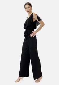Svart plisserad jumpsuit med ett lager av volangkrage, vida ben och en knytning vid midjan. Materialet verkar lätt och mjukt.