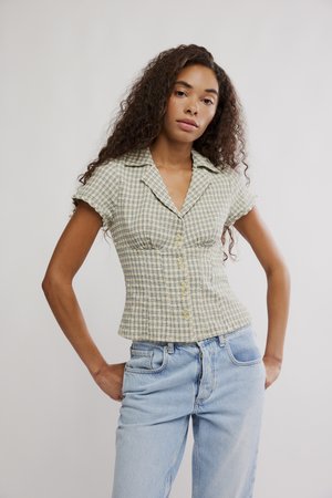 Žena s kučeravými vlasmi, oblečená v žltej károvanej blúzke s krátkym rukávom a svetlomodrých džínsach, stojaca s rukami v vreckách.