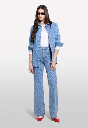 Un modèle se tient avec assurance dans une tenue en jean à motifs, accompagnée d'une chemise blanche, accessoirisée de lunettes de soleil et de talons rouges, sur un fond clair.