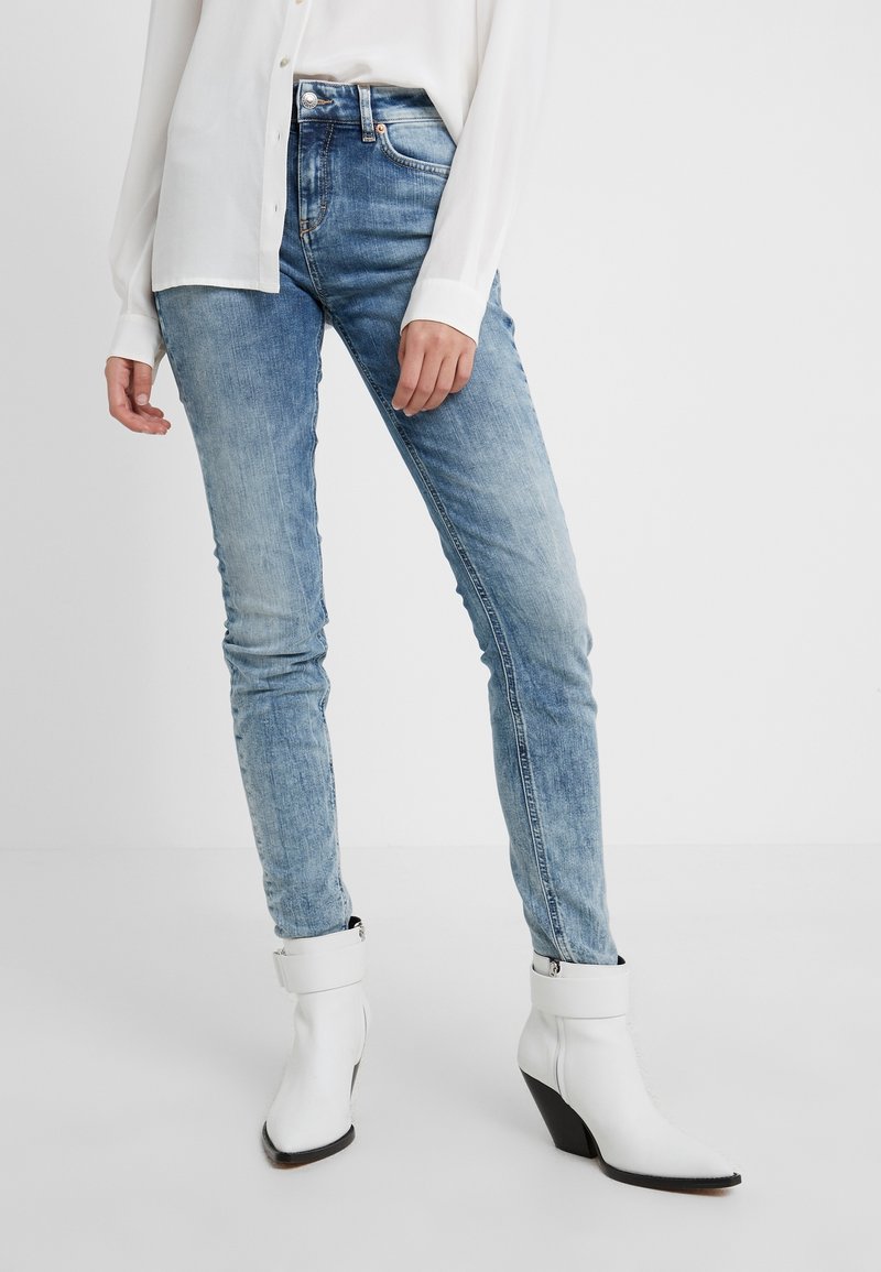 Personne portant un jean slim bleu clair et des bottines blanches à talons, associée à une chemise blanche à manches longues boutonnée.