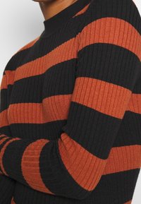 Gros plan d'une personne portant un pull en maille côtelée avec des rayures horizontales noires et orange rouille.