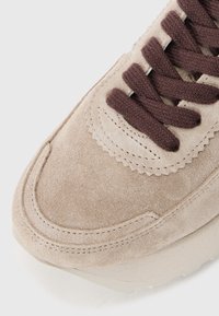 Baskets en suède beige avec lacets marron foncé, présentant un bord festonné et des détails de couture subtils. Semelle en caoutchouc avec une légère texture.