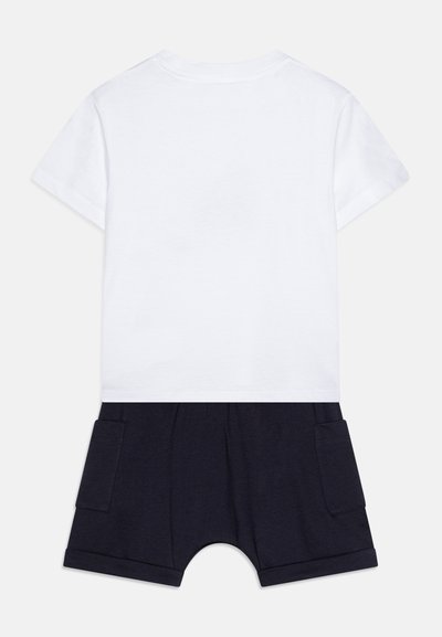 Top blanco de manga corta con parte inferior azul marino, que cuenta con dos bolsillos laterales y puños enrollados. Fabricado con tejido suave y transpirable.