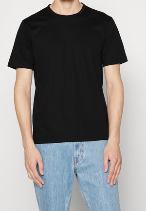 Homme portant un t-shirt noir uni à manches courtes et un jean bleu clair, debout devant un fond blanc uni.