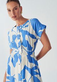 Haljina na dugme s kratkim rukavima. Ima apstraktni uzorak u plavoj i bijeloj boji, elastičan struk i glatku teksturu tkanine.