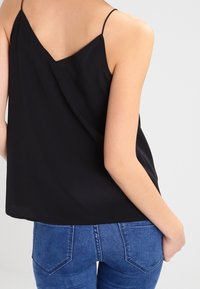 Zwarte, mouwloze top van lichtgewicht stof. Heeft een V-vormige rug en een losse pasvorm, gecombineerd met blauwe spijkerbroeken.