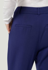 Pantalon bleu marine à texture lisse, avec deux poches arrière et une ceinture ajustée, partiellement visible avec une chemise blanche à manches longues.