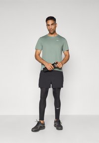 Mees rohelises Nike särgis, mustades lühikestes pükstes ja mustades retuusides, hoides käes musta veepudelit, seismas ühtlase heleda tausta ees.