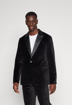 Sort fløjlsblazer med satin klap, enkelt knaplukning, to forlommer og glat tekstur. Båret over en hvid skjorte.