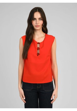 Femme aux longs cheveux foncés portant un haut rouge sans manches avec trois boutons ronds à l'avant et un jean foncé, debout devant un fond gris.