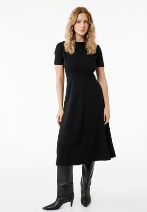 Mujer con cabello rubio ondulado que lleva un vestido midi negro ajustado de manga corta y botas negras altas con punta afilada, de pie sobre un fondo blanco.