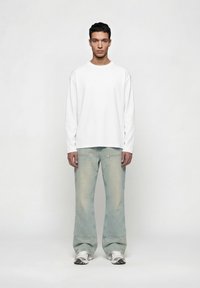 Άνδρας στέκεται μπροστά, φοράει απλό λευκό πουκάμισο με μακριά μανίκια, ξεβαμμένα τζιν με φαρδύ πόδι και λευκά αθλητικά παπούτσια σε ουδέτερο φόντο.