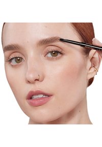 Brow pencil in hand, model applies the product to defined brown eyebrows. Skin has a smooth texture, with neutral tones and minimal makeup.