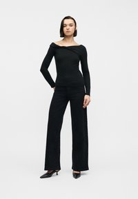 Γυναίκα που φοράει μαύρο μπλουζάκι με μακριά μανίκια χωρίς ώμους, φαρδιά μαύρη παντελόνα και μαύρες γόβες με χαμηλό τακούνι, στέκεται μπροστά σε απλό φόντο.