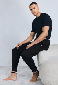 Νεαρός άνδρας με κοντά μαλλιά, ντυμένος με μαύρο μπλουζάκι και αθλητική φόρμα, καθισμένος ξυπόλητος στην μπράτσα ενός λευκού υφασμάτινου καναπέ, μπροστά σε απλό φόντο.