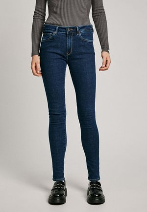 Jean skinny en denim bleu foncé, coupe taille haute, avec cinq poches et des détails de couture subtils, associés à des mocassins noirs épais.