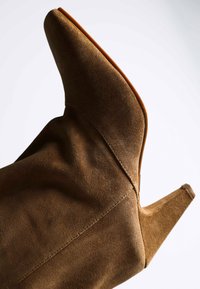 Bottine à talon haut en daim marron avec bout pointu et coutures apparentes, présentée sur un fond clair uni.