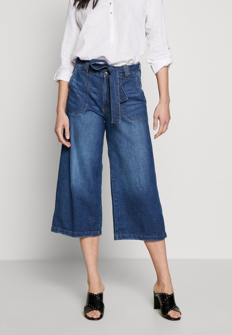 Femme portant un jean en denim taille haute, à jambes larges et coupé au-dessus de la cheville avec une ceinture, une chemise blanche à manches longues et des sandales noires ouvertes à talons.
