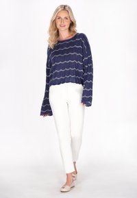 Marineblauer Strickpullover mit wellenförmigen weißen Streifen, weiten Ärmeln und einem verkürzten Saum kombiniert mit weißen, eng anliegenden Hosen und hellen Ballerinas.