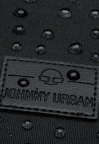 Black textured fabric with raised water droplets and an embossed label reading "JOHNNY URBAN" in a simple font. Durable and water-resistant design.