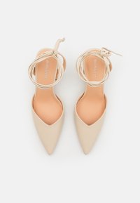 Beige høyhælte sko med spiss tådesign og omsluttende ankelbånd. Glatt tekstur og minimalistisch merkevare på innersålen.