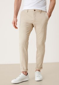 Homme portant un pantalon chino beige retroussé et des baskets blanches, debout avec une main dans la poche devant un fond uni.