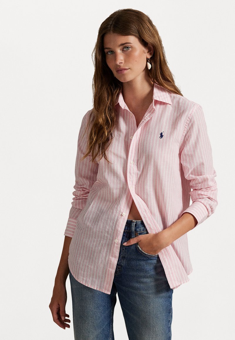 Camicia button-up a righe rosa chiaro con maniche lunghe e colletto, caratterizzata da un piccolo logo sul petto e una vestibilità rilassata. Indossata con jeans blu.