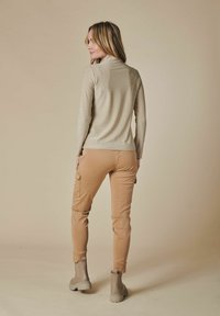 Langärmeliges, metallisch goldenes Oberteil kombiniert mit beigen Cargo-Hosen. Beigefarbene Stiefeletten mit Gummisohlen. Glatte Textur und taillierter Schnitt.