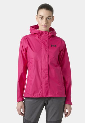 Mujer con chaqueta impermeable con capucha de color rosa brillante y logo negro en el pecho, y pantalones grises, posando frente a un fondo liso.