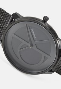 Svart mesh klokke med en matt grå rund urskive som har minimalistiske timeindikatorer og Calvin Klein-logo. Elegant kasse med en sidekrone.