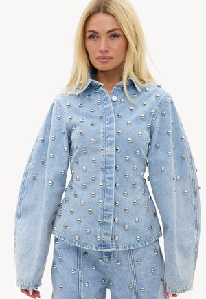 Femme blonde portant une veste et un pantalon en denim bleu clair avec des clous argentés répartis uniformément, debout devant un fond blanc uni.