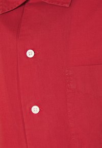 Chemise rouge en tissu lisse avec une patte de boutonnage à boutons blancs, un col pointu et une poche poitrine sur le côté gauche.