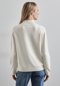 Crème kleurige gebreide trui met een geribbelde kraag, relaxte pasvorm, lange mouwen en een gestructureerde afwerking. Gecombineerd met blauwe spijkerbroek.