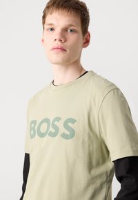 Gaiši zaļš kokvilnas T-krekls ar izteiktu "BOSS" logo tumši zaļā krāsā. Vilkts virs melna garām piedurknēm krekla ar vienkāršu dizainu.