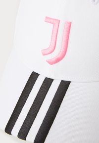 Balts beisbola cepure ar rozā izšūtu logotipu un melniem svītrainiem akcentiem uz mala. Izgatavota no teksturēta, viegla auduma.
