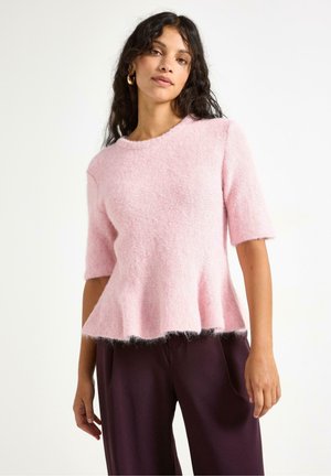 Maglione rosa chiaro a maniche corte con orlo svasato e texture morbida e pelosa. Abbinato a pantaloni larghi di colore viola scuro.