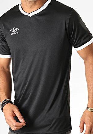 Uomo che indossa una maglietta sportiva nera a maniche corte con bordino bianco, logo Umbro sul petto, braccialetti neri con perline al polso sinistro e orologio d'oro al polso destro.