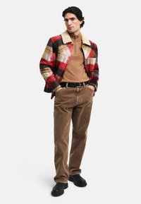 Chaqueta de lana a cuadros en rojo, negro y beige, con un collar de piel de oveja. Suéter de cuello con cremallera en color marrón debajo. Pantalones de pana marrón y zapatos negros.